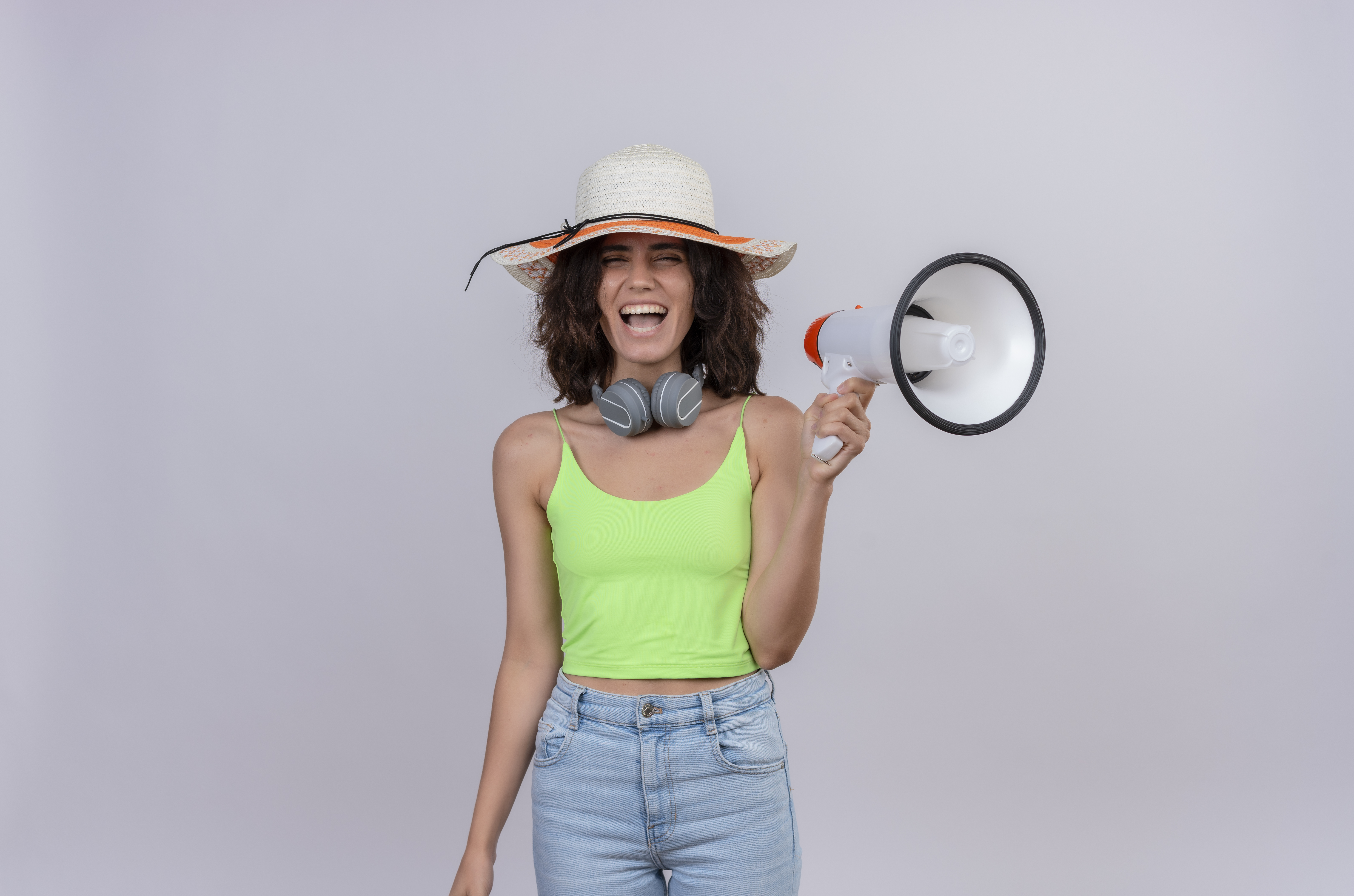 a happy young woman with short hair in green crop top in headphones wearing sun hat smiling and holding megaphone on a white background